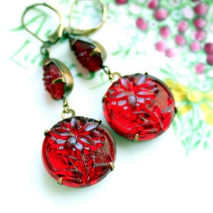 May include: A pair of drop earrings featuring round, deep red glass cabochons with a molded floral design. Each earring has a smaller, teardrop-shaped red glass accent above the circular piece, all set in an antique brass-toned metal frame.