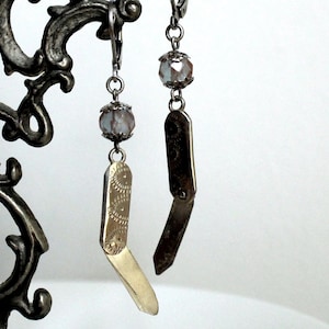 May include: A pair of dangle earrings featuring silver-toned leverback hooks, each adorned with a faceted, pale iridescent bead held by metal caps. Below each bead hangs a small, hinged metal charm shaped like a folding pocket knife.
