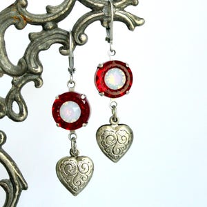 May include: A pair of drop earrings featuring a round, faceted red crystal with an iridescent white center, suspended above a small, silver-toned heart charm with an embossed scrollwork pattern, attached to silver-toned lever-back ear wires.