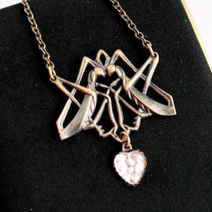 May include: A bronze-toned metal necklace featuring a pendant shaped like two mirrored insects facing each other, connected by a thin chain. A small, heart-shaped charm with a textured, pearlescent white inlay hangs from the center of the insect design.