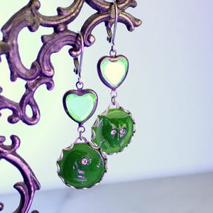 May include: A pair of drop earrings featuring a light green heart-shaped charm attached to a circular dark green disc with a molded cat face design. The earrings are constructed with silver-toned metal settings and include small clear crystal accents.