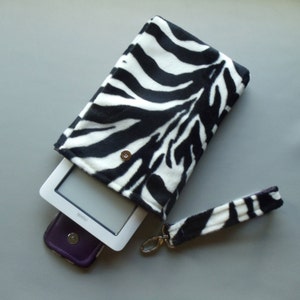 May include: A black and white zebra print ebook reader case with a matching wrist strap. The case is open, revealing a white ebook reader and a purple phone case. The case has a magnetic snap closure.