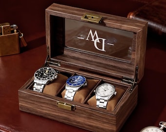 Puede incluir: Una caja de reloj de madera abierta que muestra tres relojes de pulsera con correas plateadas y esferas de diferentes colores, colocados sobre cojines marrones. También se ve un encendedor dorado y un reloj de bolsillo.
