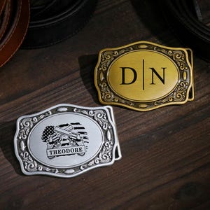 Puede incluir: Dos hebillas decorativas para cinturón. Una es dorada con las iniciales "D | N" en negro. La otra es plateada, con un gráfico de revólveres cruzados, una bandera estadounidense y el nombre "THEODORE".