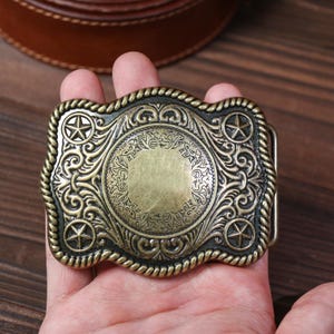 May include: A vintage brass-toned belt buckle with a Western design. The rectangular buckle has a central circular pattern with detailed scrollwork, four star motifs, and a rope-style edge. The buckle is held in a hand.