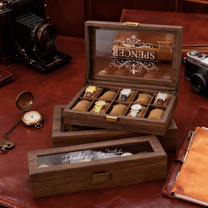 May include: A wooden watch display case with a glass top, holding several wristwatches. The case is open, revealing the watches on brown cushions. The top of the case is engraved with the name "Spencer". A smaller case below reads "Daddy's Watch".