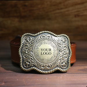 May include: An antique-style belt buckle with a bronze finish. The buckle has a central circular design with the text "YOUR LOGO", surrounded by ornate scrollwork and star accents. It is attached to a brown leather belt. The buckle is approximately 10 cm wide.