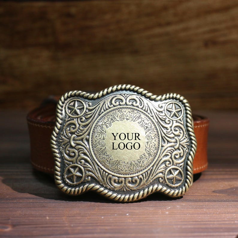 May include: An antique-style belt buckle with a bronze finish. The buckle has a central circular design with the text "YOUR LOGO", surrounded by ornate scrollwork and star accents. It is attached to a brown leather belt. The buckle is approximately 10 cm wide.