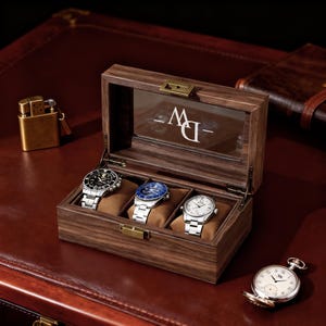 May include: A wooden watch box with three watches inside, each resting on a brown cushion. The box has a glass top and a metal clasp. A gold lighter and a pocket watch are also visible.