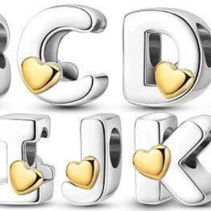 May include: Silver alphabet charms from A to M, each featuring a gold heart detail. These charms are designed for bracelets or necklaces, providing a personalised element. The letters are in a bold, rounded font.