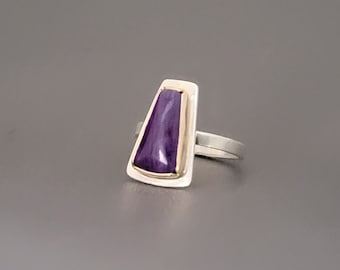 May include: A silver ring featuring a purple stone set in a gold bezel. The ring has a simple, modern design.