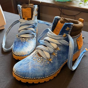 Può includere: Un paio di stivaletti in velluto azzurro con linguetta in pelle color cuoio con logo Timberland, ampi lacci in tessuto azzurro e una fila di strass blu lungo la base e gli occhielli. Gli stivali hanno una suola carrarmato in gomma color cuoio.