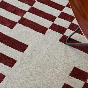 May include: A close-up of a rug with a geometric pattern. The rug features alternating white and burgundy rectangular shapes. A brown leather chair is partially visible in the upper right corner, with a black metal frame.
