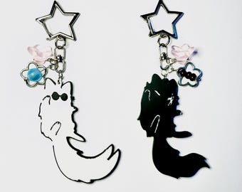 May include: Two keychains featuring star-shaped silver clasps. One keychain has a white fox charm with black sunglasses, a pink flower bead, and a blue bead. The other has a black fox charm, a pink flower bead, and three small black beads.