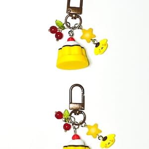 May include: Two keychains with a yellow dessert charm, red cherry accents, and a yellow star and character charm. Each keychain has a bronze-colored clasp and ring. The charms are attached with silver-colored rings.