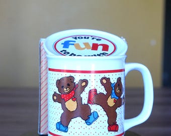 May include: White ceramic mug with a lid, featuring two brown teddy bears dancing. The mug has a red and white polka dot pattern and a red stripe. The lid says "You're fun to be with".