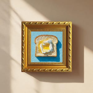 Puede incluir: Pintura enmarcada de una rebanada de pan tostado con un huevo frito. La obra de arte está sobre un fondo azul claro y enmarcada en un marco dorado ornamentado. Es una naturaleza muerta.