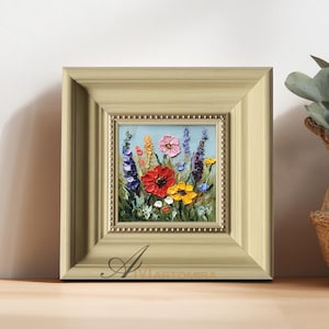Puede incluir: Pintura al óleo enmarcada de coloridas flores silvestres. La obra de arte presenta una vibrante variedad de flores rojas, rosas, amarillas y moradas sobre un fondo azul claro. La pintura está enmarcada en un marco beige claro con un borde interior con cuentas.