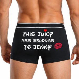 Könnte beinhalten: Schwarze Boxershorts mit weißem Text "THIS JUICY ASS BELONGS TO JENNY". Ein rotes Herz und ein roter Kussmund sind ebenfalls auf der Hose abgebildet. Der Bund hat einen weißen und grauen Streifen.