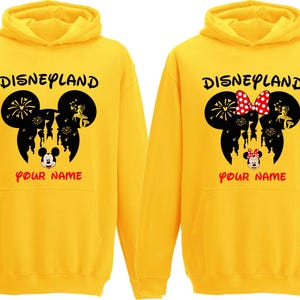 以下が含まれることがあります： 「Disneyland」の文字と黒いミッキーマウスの頭のシルエットがプリントされた2つの黄色のパーカー。1つはミッキーマウスのデザイン、もう1つは赤い水玉模様のリボンが付いたミニーマウスのデザインです。「YOUR NAME」の文字が下にあります。