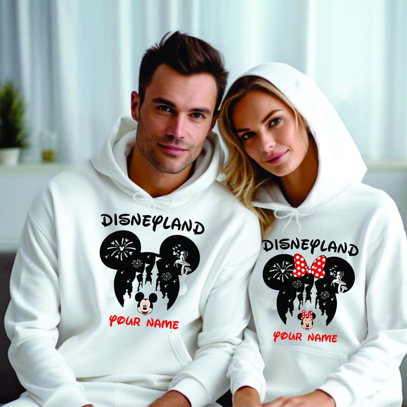 以下が含まれることがあります： 白のフード付きスウェットシャツ。黒で「Disneyland」とプリントされ、ミッキーマウスのシルエットデザインが施されています。デザインには花火と城が含まれています。「Your Name」の文字は赤でプリントされています。