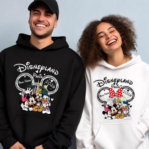 Puede incluir: Sudaderas con capucha negras y blancas de Disneyland Par&iacute;s 2026. El dise&ntilde;o presenta a Mickey y Minnie Mouse, el Pato Donald y Goofy. El texto "Disneyland Par&iacute;s" est&aacute; arqueado sobre los personajes. El dise&ntilde;o incluye el texto "Your Name" y el a&ntilde;o "2026".