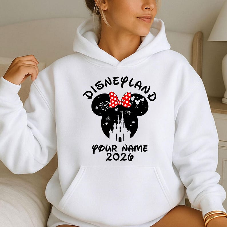 May include: White hooded sweatshirt featuring a black Minnie Mouse silhouette with a red and white polka dot bow. The word "Disneyland" is arched above the silhouette, with "Your Name 2026" printed below in black.