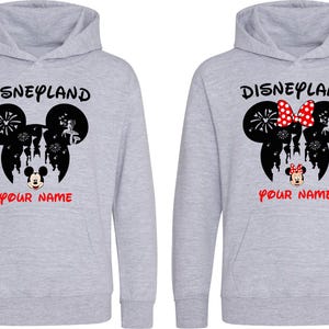 以下が含まれることがあります： 「Disneyland」の文字と黒いミッキーマウスの頭のデザインが施された2つのグレーのパーカー。1つのパーカーには、白のドットが付いた赤いリボンが付いています。「YOUR NAME」の文字が赤色で下に印刷されています。