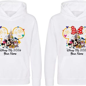 May include: Two white hooded sweatshirts featuring a graphic of Mickey Mouse, Minnie Mouse, Goofy, Donald Duck, and Pluto in front of a castle. The design includes a yellow Mickey-shaped outline, fireworks, and the text Disney Trip 2026 Your Name.