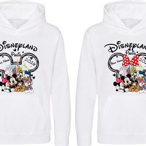 Puede incluir: Sudaderas con capucha blancas con un dise&ntilde;o de Disneyland Par&iacute;s. El gr&aacute;fico incluye a Mickey Mouse, Minnie Mouse, el Pato Donald, Goofy y Pluto. El dise&ntilde;o presenta el texto "Disneyland Par&iacute;s 2020" y un espacio para un nombre. Una sudadera tiene un lazo rojo de lunares.