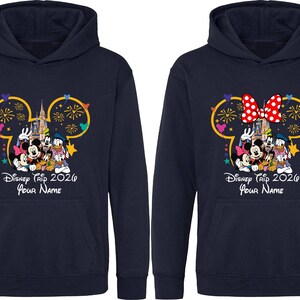 May include: Two navy blue hooded sweatshirts featuring a graphic of Disney characters in front of a castle with fireworks inside a Mickey Mouse silhouette outline. The text reads Disney Trip 2026 and Your Name, with one hoodie featuring a red polka dot bow.
