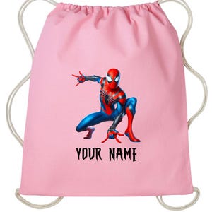 May include: A light pink drawstring backpack featuring a colorful illustration of Spider-Man in a crouching pose. The superhero is in a red and blue suit. The text "YOUR NAME" is printed in black below the image.