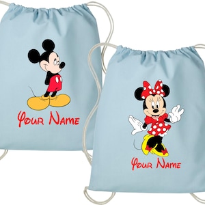 Op de afbeelding: Twee lichtblauwe trekkoordtassen met Mickey Mouse en Minnie Mouse. Mickey draagt een rood shirt en gele schoenen. Minnie draagt een rode polkadotjurk en strik. De tekst "Your Name" staat eronder.