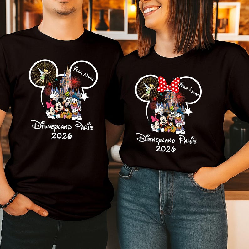 May include: Black t-shirts featuring a Disney theme. The design includes Mickey and Minnie Mouse, a castle, fireworks, and the text "Disneyland Paris 2026". One shirt has a red bow, and both have a speech bubble with "Your Name".