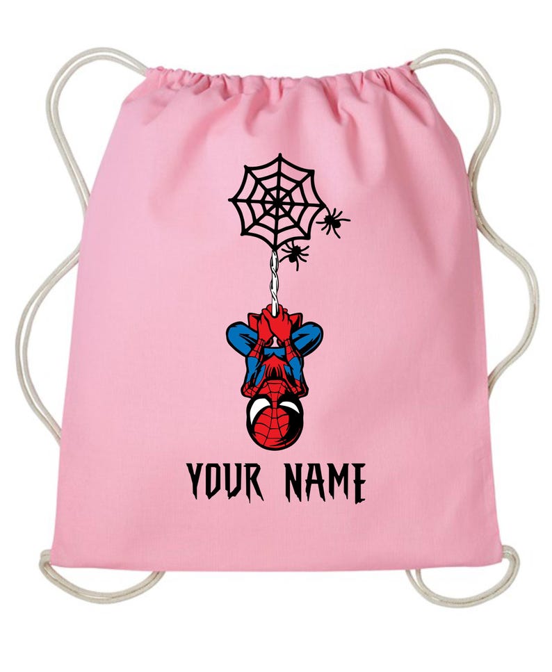 Puede incluir: Mochila con cord&oacute;n rosa con un dise&ntilde;o de Spider-Man. El dise&ntilde;o incluye una telara&ntilde;a negra, ara&ntilde;as y Spider-Man colgado boca abajo. El texto "YOUR NAME" est&aacute; impreso en negro.