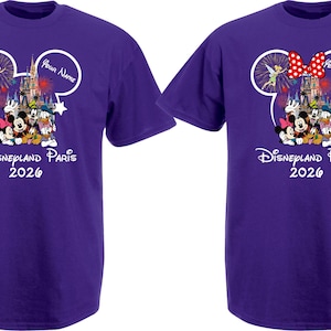 May include: Two purple t-shirts with a Disney theme. Each shirt features a Mickey Mouse head outline with a castle, fireworks, and Disney characters. The text "Disneyland Paris 2026" is printed below the graphic. One shirt has a red bow.