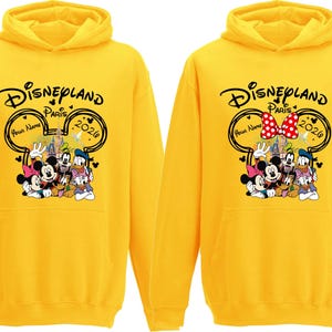 Puede incluir: Dos sudaderas con capucha amarillas con el logotipo de Disneyland Paris, con Mickey Mouse, Minnie Mouse, el Pato Donald, Goofy y Pluto. Las sudaderas tienen el texto "Disneyland Paris 2026" y un espacio para un nombre.