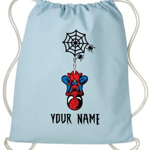 Puede incluir: Mochila con cord&oacute;n azul claro con un dise&ntilde;o de Spider-Man. El dise&ntilde;o incluye una telara&ntilde;a negra, dos ara&ntilde;as y Spider-Man colgado boca abajo. El texto "YOUR NAME" est&aacute; impreso en negro debajo.