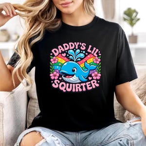 May include: A black t-shirt featuring a colourful graphic of a blue whale spouting water, surrounded by a rainbow, pink hibiscus flowers, and small hearts. The text DADDY'S LIL SQUIRTER is printed in pink capital letters around the central illustration.