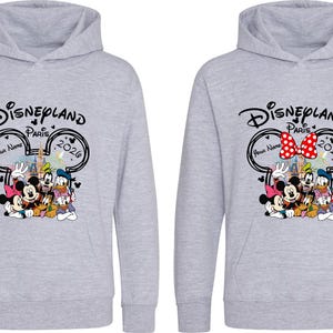 Puede incluir: Dos sudaderas con capucha de color gris claro con un gr&aacute;fico de Disneyland Paris 2020 con Mickey Mouse, Minnie Mouse, Donald Duck y Goofy. Una sudadera con capucha tiene un lazo rojo y blanco. La otra tiene un lazo negro.