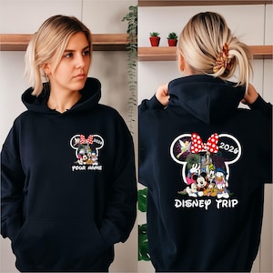 Könnte beinhalten: Marineblauer Kapuzenpullover mit Minnie Mouse-Design, roter Schleife und der Jahreszahl "2026". Auf der Vorderseite steht "YOUR NAME" unter der Grafik. Auf der Rückseite steht "DISNEY TRIP" und eine farbenfrohe Disney-Charakter-Illustration.