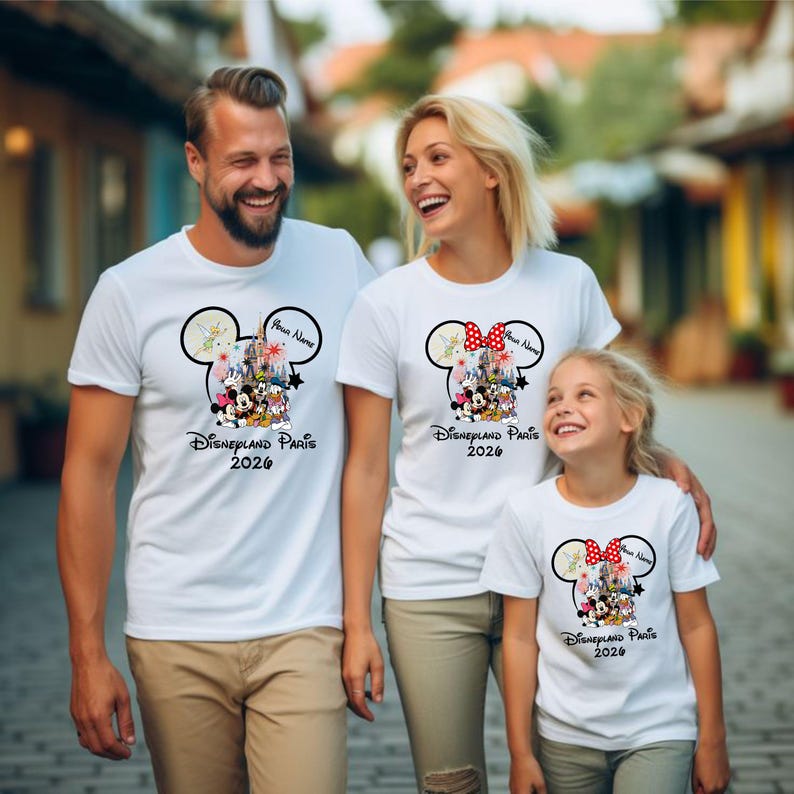May include: Three people wearing matching white t-shirts with a Disneyland Paris 2026 design. The design features a Mickey Mouse head with a castle, characters, and a name bubble. The shirts are for a family.