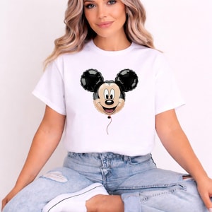 May include: White t-shirt featuring a Mickey Mouse balloon graphic. The balloon has black ears, a tan face, and a red string. The shirt is made of a soft material and is suitable for casual wear.