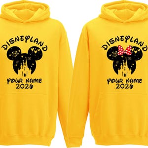 May include: Two yellow Disneyland hoodies. One features a black Mickey Mouse head design with fireworks, a castle, and the text "Disneyland" and "Your Name 2026." The other has a Minnie Mouse head with a red bow.