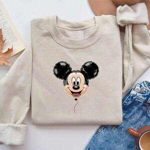 May include: A beige sweatshirt featuring a Mickey Mouse balloon design. The balloon has black ears and a smiling face. The sweatshirt is folded, with a pair of blue jeans, a hat, and a cup of coffee in the background.