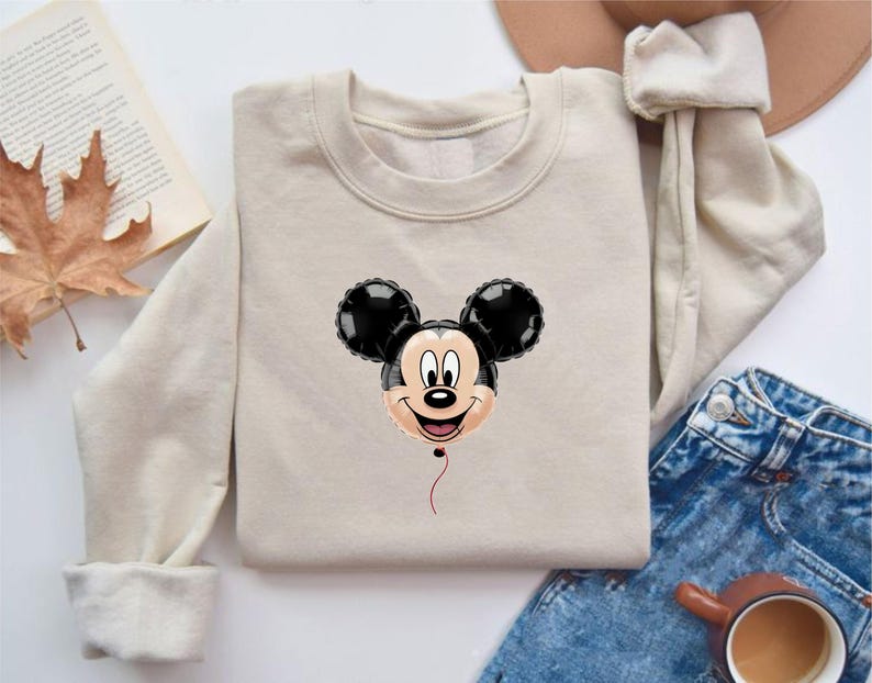 May include: A beige sweatshirt featuring a Mickey Mouse balloon design. The balloon has black ears and a smiling face. The sweatshirt is folded, with a pair of blue jeans, a hat, and a cup of coffee in the background.