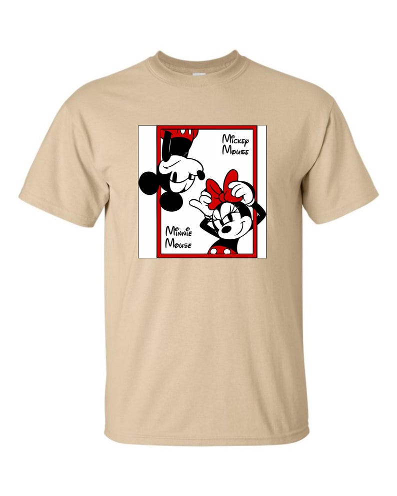 May include: A tan t-shirt featuring a graphic of Mickey Mouse and Minnie Mouse. The graphic is in a red rectangular frame with the words "Mickey Mouse" and "Minnie Mouse" in black text. Mickey is upside down, and Minnie is right-side up.