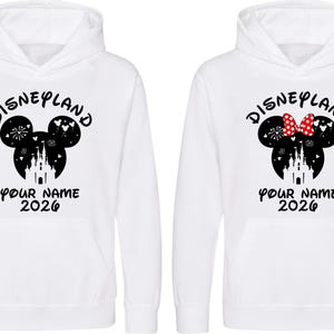 May include: Two white hooded sweatshirts with a Disneyland design. One features a Mickey Mouse silhouette with fireworks, a castle, and the text "YOUR NAME 2026". The other has a Minnie Mouse silhouette with a red bow.