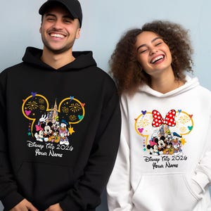 May include: A black hoodie and a white hoodie, each featuring a graphic of Mickey Mouse, Minnie Mouse, Goofy, and Donald Duck in front of a castle. The design includes a Mickey-shaped outline, fireworks, and the text Disney Trip 2026 Your Name.