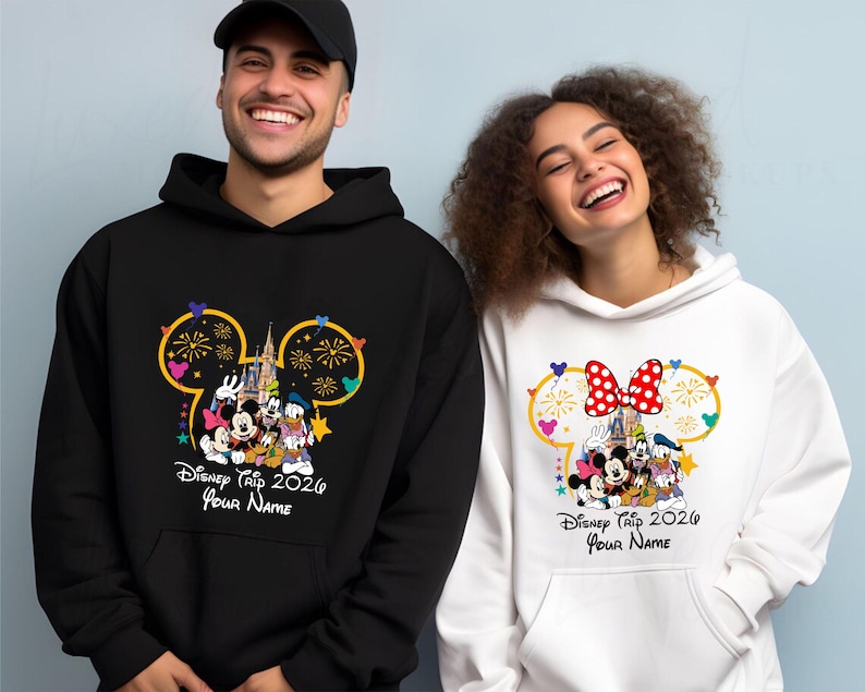 May include: A black hoodie and a white hoodie, each featuring a graphic of Mickey Mouse, Minnie Mouse, Goofy, and Donald Duck in front of a castle. The design includes a Mickey-shaped outline, fireworks, and the text Disney Trip 2026 Your Name.